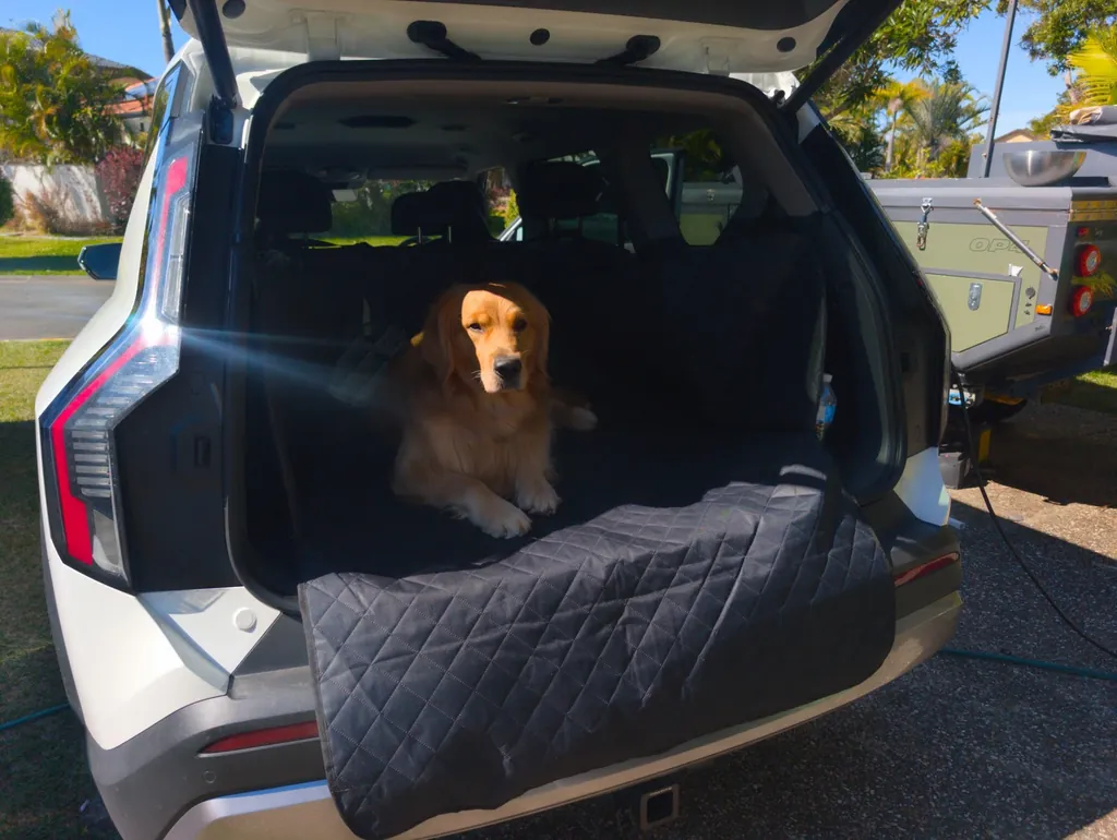 A golden retriever lying in the boot of a white EV9 on a black quilted liner, with the liner’s flap folded down over the bumper.